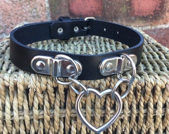 Puede incluir: Choker de cuero negro con herrajes plateados y un dije en forma de corazón. El choker tiene dos hebillas plateadas y una cadena con un dije en forma de corazón.
