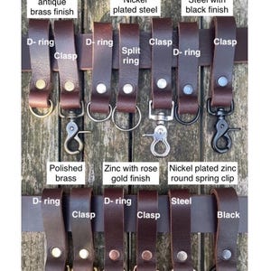 May include: A collection of brown buffalo leather belt loops featuring various metal hardware attachments including D-rings, clasps, split rings, and spring clips in finishes such as antique brass, nickel, black, polished brass, and rose gold.