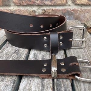 Peut inclure: Deux ceintures en cuir marron foncé avec boucles argentées. Les ceintures sont en cuir épais et comportent plusieurs trous de réglage. Les boucles sont carrées et les ceintures sont présentées sur une surface en bois.