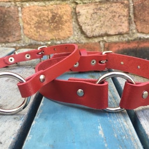 May include: Two red leather collars with silver metal rings. The collars are on a wooden surface.