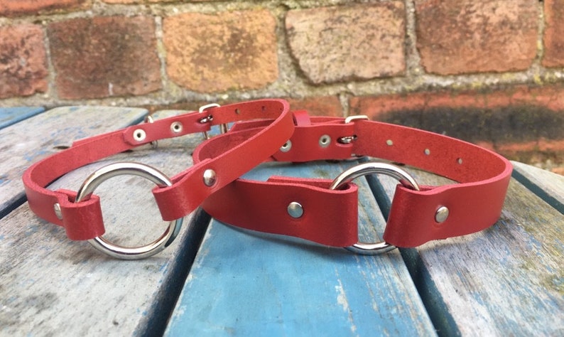 May include: Two red leather collars with silver metal rings. The collars are on a wooden surface.