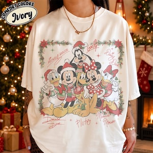 May include: An ivory t-shirt featuring a festive graphic of Disney characters including Mickey Mouse, Minnie Mouse, Donald Duck, Daisy Duck, Goofy, and Pluto wearing holiday attire. The design is framed by a wreath with signatures and text.