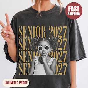 Può includere: Una t-shirt grigio antracite con un motivo di testo dorato ripetuto che recita SENIOR 2027. La grafica centrale mostra una fotografia in bianco e nero di un bambino con le codine che indossa occhiali da sole rotondi a forma di fiore.