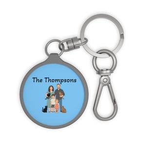 May include: A round metal keychain with a light blue background featuring a custom illustration of a family of four and two cats. The text "The Thompsons" is printed in black above the family. The keychain includes a silver ring and a lobster clasp.