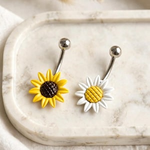 May include: Two curved barbell body jewelry pieces featuring flower designs. One piece has a yellow sunflower with a dark brown center, and the other has a white daisy with a yellow center. Both have a polished silver-tone metal ball at the top.
