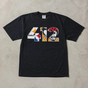 May include: A black short-sleeve t-shirt featuring the large white numbers 412 on the chest. The number 4 contains the Pittsburgh Steelers logo, the number 1 features a penguin graphic, and the number 2 displays a pirate character with a red bandana.