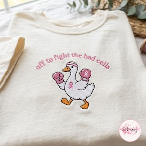 May include: A cream-colored t-shirt featuring an embroidered graphic of a white duck wearing pink boxing gloves and a pink headband. The duck has a pink ribbon on its chest. The text "off to fight the bad cells" is embroidered in pink above the duck.