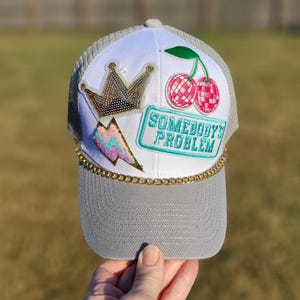 May include: A white and gray trucker hat featuring a gold sequin crown patch, a pink and blue lightning bolt patch, a pair of pink disco ball cherries, and a teal rectangular patch with the text SOMEBODY'S PROBLEM, accented by a gold chain.