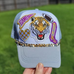 May include: A light blue trucker hat with a white front panel featuring a tiger face, football, heart, and cowboy boot patches. The hat has the word "GAMEDAY" and gold sequins along the brim.