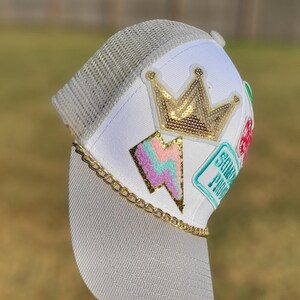May include: A white and grey trucker hat featuring a gold sequined crown patch, a pastel lightning bolt patch, and a teal rectangular patch with text. A gold-tone metal chain trim sits along the base of the front panel above the grey brim.