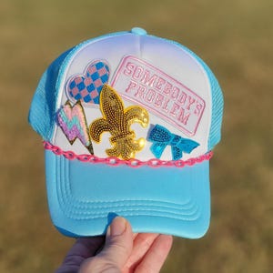 May include: A light blue trucker hat with a white front panel featuring a pink rectangular patch that reads SOMEBODY'S PROBLEM. The hat is decorated with a checkered heart, a lightning bolt, a gold sequined fleur-de-lis, and a blue bow.