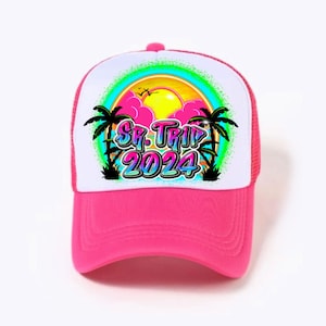 May include: A pink trucker hat with a white front panel featuring a colorful graphic. The graphic includes palm trees, a sunset, and the text "Sr. Trip 2024" in a vibrant, multi-colored font. The hat has a pink brim and mesh backing.