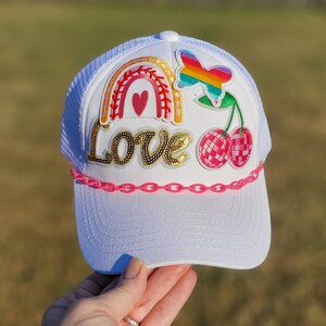 May include: White trucker hat with a rainbow, butterfly, and cherry design. The word "Love" is spelled out in gold sequins. A pink chain detail accents the brim. The hat is a fun and colorful accessory.