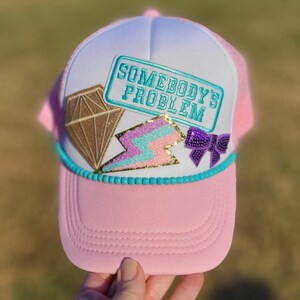 May include: Pink trucker hat with a white front panel and a pink brim. The hat features the text "SOMEBODY'S PROBLEM" in a teal rectangle, a gold diamond, a lightning bolt, and a purple bow.