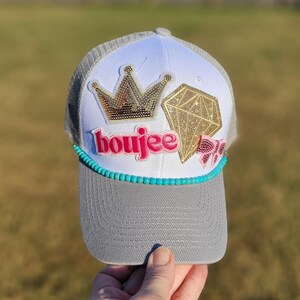 May include: A gray and white trucker hat with a curved brim. The hat features a gold crown and diamond applique, the word "boujee" in pink, and a turquoise beaded band. The hat is held by a hand.