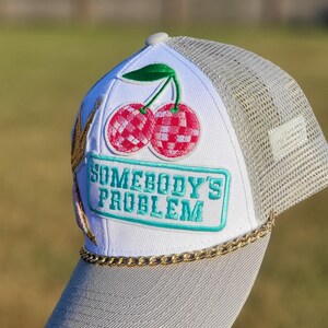May include: A white trucker hat with a grey mesh back and brim. The front features an embroidered patch of two pink disco ball cherries with a green leaf. Below is a rectangular teal patch with the text SOMEBODY'S PROBLEM and a gold chain trim.