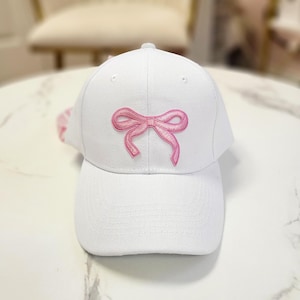 May include: White baseball cap with a pink embroidered bow on the front. The cap has a curved brim and a structured crown. The bow is a delicate, feminine design.