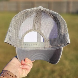 May include: A light grey trucker hat featuring a mesh back panel and a solid front panel. The cap includes an adjustable plastic snapback closure at the rear, a structured crown, and a curved brim, all rendered in a consistent monochromatic grey tone.