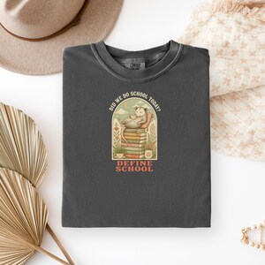 May include: A charcoal gray crewneck t-shirt featuring a graphic of an opossum resting on a stack of books. The text above the opossum reads DID WE DO SCHOOL TODAY? and the text below the stack of books reads DEFINE SCHOOL.