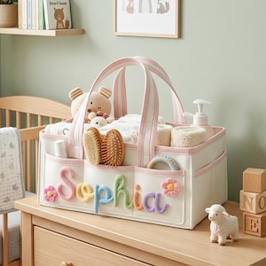 May include: A white rectangular fabric storage caddy with pink gingham trim and handles. The front features the name Sophia in colorful, textured yarn letters, accented by two small crocheted flowers. It is filled with baby care items and brushes.