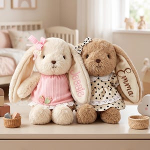 May include: Two plush stuffed rabbits with long ears. The cream-colored rabbit wears a pink dress with strawberry accents and a gingham bow. The brown rabbit wears a cream dress with black polka dots and a bow, featuring the names Jessie and Emma.