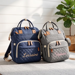 May include: Two structured backpacks with top handles and adjustable straps. The navy blue bag features the name Lorraine embroidered in gold, while the grey bag features the name Stephanie embroidered in pink. Both have quilted front pockets.
