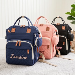 Pode incluir: Três mochilas estruturadas em azul-marinho, rosa-claro e preto. Cada bolsa possui um bolso frontal acolchoado, alças superiores, alças de ombro ajustáveis e uma pequena bolsa circular presa na lateral. Os nomes Lorraine, Janet e Renee estão bordados nos bolsos frontais em letra cursiva.