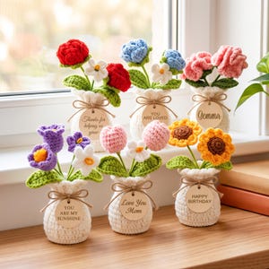 Könnte beinhalten: Sechs gehäkelte Blumenarrangements in kleinen, weißen, strukturierten Vasen. Jede Vase hat ein rundes Holzschild mit Text, darunter Thanks for helping us grow, You are loved, Gemma, You are my sunshine, Love You Mom und Happy Birthday.