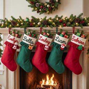 May include: Five Christmas stockings in alternating red and green colors, each featuring a white cuff with a ruffled edge and a personalized name embroidered in cursive: Evelyn, Bella, Lesley, Liam, and Sabrina. Each stocking holds small gift boxes.