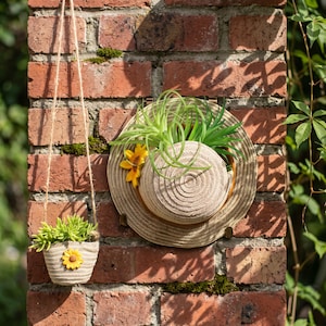 Op de afbeelding: Een beige strooien hoed met een ronde rand en een geweven kroon, versierd met een gele zonnebloem en groene luchtplanten. Daarnaast hangt een kleine, bijpassende beige stoffen plantenbak met een groene vetplant en een gele zonnebloem.