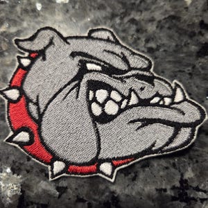 Puede incluir: Un parche bordado que muestra la cabeza de un bulldog gris con expresión gruñona, dientes blancos al descubierto y un collar rojo con pinchos. El diseño está delineado con hilo negro y un borde blanco, mostrando costuras detalladas en la imagen.