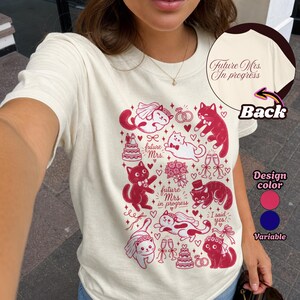 Puede incluir: Una camiseta color crema con un estampado gráfico rojo de gatos con ropa de boda, pasteles, anillos y copas de champán. El frente incluye el texto "future Mrs. in progress" y "I said yes!" mientras que la parte posterior muestra "Future Mrs. In progress."