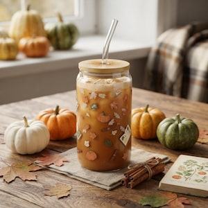 Puede incluir: Un vaso de cristal transparente lleno de café helado, con un estampado de pequeñas calabazas, hojas de otoño y libros. El vaso tiene una tapa de bambú y una pajita de cristal. La bebida es de color marrón claro con cubitos de hielo.