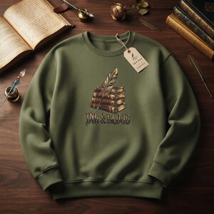 Puede incluir: Una sudadera de cuello redondo color verde oliva con un gráfico de una pila de cuatro libros encuadernados en cuero con una pluma dorada encima. Debajo de los libros, el texto "INK & BLOOD" aparece en una fuente estilizada. Tiene una etiqueta de papel beige.