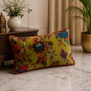 Peut inclure: Un coussin rectangulaire en velours vert olive orné d'un motif floral aux couleurs vives, incluant des tons rouge profond, rose vif et bleu foncé. Le coussin est bordé d'un fin passepoil rouge contrastant sur tout son pourtour.