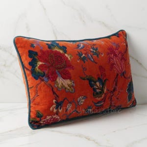 May include: A rectangular throw pillow featuring a vibrant orange base with a multi-colored floral pattern in shades of red, blue, and green. The pillow is finished with a dark teal piped edge and includes the text Theshanazstore in the center.