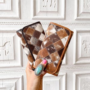 May include: Two rectangular wallets featuring a patchwork design of cowhide in shades of brown, white, and black. The wallet on the left has dark brown trim, while the wallet on the right has tan trim. Both wallets have a flap closure.