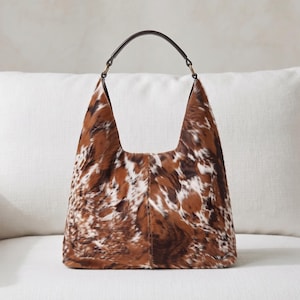 May include: A hobo-style handbag featuring a brown, white, and black cowhide pattern with a textured fur finish. The bag has a single dark brown leather handle attached with metal rings, a wide opening, and a vertical seam down the center.
