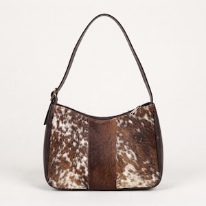 May include: A shoulder bag featuring a brown and white cowhide pattern on the front panels with a solid dark brown leather center stripe. The bag includes a single dark brown leather strap, a top zipper closure, and dark brown leather sides.
