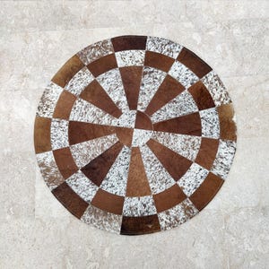 May include: A circular rug constructed from alternating segments of brown leather and white and brown speckled cowhide. The pattern radiates from a central circular piece, creating a geometric, multi-paneled design with a textured finish.