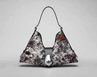 May include: A triangular hobo bag featuring a black and white cowhide pattern with a smooth black leather strap and a central vertical black leather tab secured by a large, polished, silver-toned oval metal clasp at the bottom center.