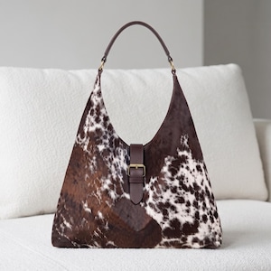 May include: A hobo-style handbag featuring a brown and white cowhide pattern with a smooth dark brown leather strap and a central vertical leather tab closure secured by a brass-toned metal buckle, finished with a single matching leather handle.