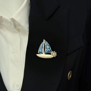 May include: A gold-toned sailboat brooch featuring blue patterned sails and a small ship's wheel attached to the side. The sailboat has a smooth, curved hull and is pinned to the lapel of a black blazer worn over a white collared shirt.