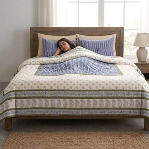 May include: A bed featuring a cream-coloured quilt with a repeating olive green paisley border and a central blue and white chevron patterned section. Two matching blue and white chevron pillows are placed against a wooden headboard.
