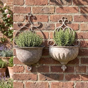 May include: Two wall-mounted metal planters with a weathered, rustic finish. Each planter features an ornate, scrolled metal backplate and a semi-circular basin. One basin has a textured rim, while the other has vertical fluted detailing.