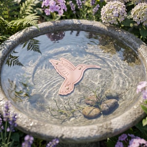 Może przedstawiać: Płaski obiekt w kolorze jasnoróżowym w kształcie kolibra z metalowymi nitami w kolorze złotym, z nadrukowanym napisem "100% Pure Copper" oraz "Bird Bath", spoczywający zanurzony w okrągłej kamiennej poidełku dla ptaków wypełnionym czystą wodą.