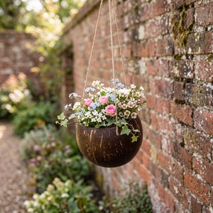 May include: A round, dark brown coconut shell hanging planter suspended by thin jute twine. The planter is filled with a variety of small artificial flowers, including pink roses, light blue forget-me-nots, white blossoms, and green ivy leaves.