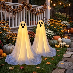 May include: Two illuminated ghost decorations made of white fabric, each featuring a printed black face with two oval eyes and an oval mouth. The figures emit a warm internal glow and are positioned upright on a grassy lawn surface.