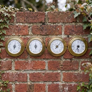 May include: Four circular brass-rimmed instruments mounted in a row. From left to right, they include a thermometer with a white face, a hygrometer, a barometer with weather icons, and a tide clock. Each features a black needle and markings.