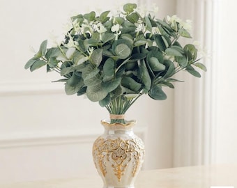 Op de afbeelding: Een boeket van kunstmatige groene eucalyptusbladeren met kleine witte bloemen in een parelmoerwitte vaas met sierlijke goudkleurige reliëfdetails rond de buik en een uitlopende rand, staand op een glad oppervlak.